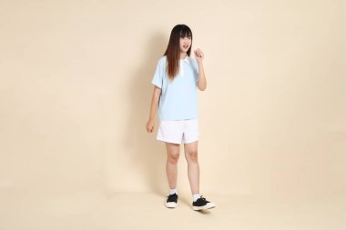 Açık mavi polo tişörtlü, beyaz şortlu, bej arka planda yürüyen genç Asyalı bir kadın. Üniversite öğrencisi, Gen Z, Summer