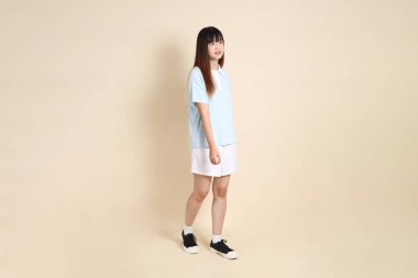 Açık mavi polo tişörtlü, beyaz şortlu, bej arka planda yürüyen genç Asyalı bir kadın. Üniversite öğrencisi, Gen Z, Summer