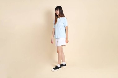 Açık mavi polo tişörtlü, beyaz şortlu, bej arka planda yürüyen genç Asyalı bir kadın. Üniversite öğrencisi, Gen Z, Summer