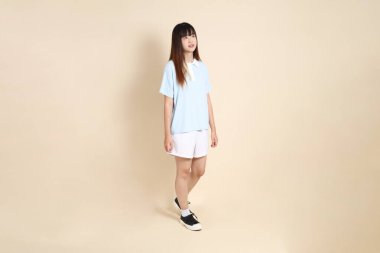 Açık mavi polo tişörtlü, beyaz şortlu, bej arka planda yürüyen genç Asyalı bir kadın. Üniversite öğrencisi, Gen Z, Summer