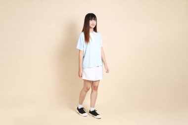 Açık mavi polo tişörtlü, beyaz şortlu, bej arka planda yürüyen genç Asyalı bir kadın. Üniversite öğrencisi, Gen Z, Summer