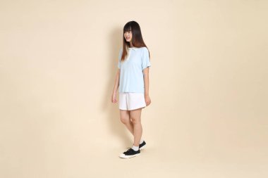 Açık mavi polo tişörtlü, beyaz şortlu, bej arka planda yürüyen genç Asyalı bir kadın. Üniversite öğrencisi, Gen Z, Summer