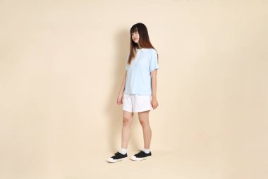 Açık mavi polo tişörtlü, beyaz şortlu, bej arka planda yürüyen genç Asyalı bir kadın. Üniversite öğrencisi, Gen Z, Summer
