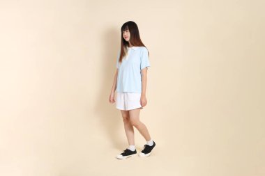 Açık mavi polo tişörtlü, beyaz şortlu, bej arka planda yürüyen genç Asyalı bir kadın. Üniversite öğrencisi, Gen Z, Summer