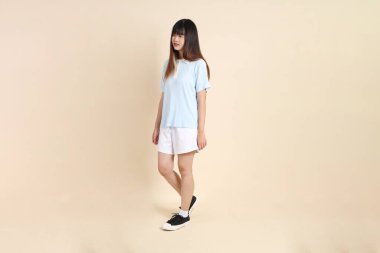 Açık mavi polo tişörtlü, beyaz şortlu, bej arka planda yürüyen genç Asyalı bir kadın. Üniversite öğrencisi, Gen Z, Summer