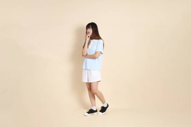 Açık mavi polo tişörtlü, beyaz şortlu, bej arka planda yürüyen genç Asyalı bir kadın. Üniversite öğrencisi, Gen Z, Summer