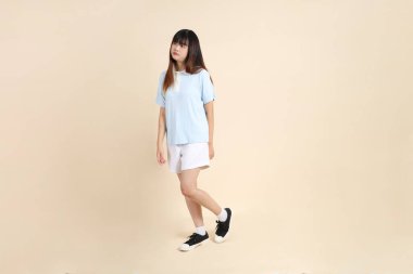Açık mavi polo tişörtlü, beyaz şortlu, bej arka planda yürüyen genç Asyalı bir kadın. Üniversite öğrencisi, Gen Z, Summer