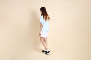 Açık mavi polo tişörtlü, beyaz şortlu, bej arka planda yürüyen genç Asyalı bir kadın. Üniversite öğrencisi, Gen Z, Summer