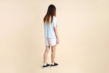 Açık mavi polo tişörtlü, beyaz şortlu, bej arka planda yürüyen genç Asyalı bir kadın. Üniversite öğrencisi, Gen Z, Summer