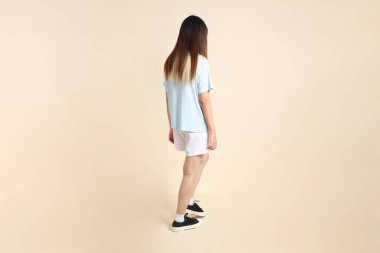 Açık mavi polo tişörtlü, beyaz şortlu, bej arka planda yürüyen genç Asyalı bir kadın. Üniversite öğrencisi, Gen Z, Summer