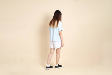 Açık mavi polo tişörtlü, beyaz şortlu, bej arka planda yürüyen genç Asyalı bir kadın. Üniversite öğrencisi, Gen Z, Summer