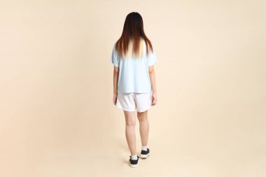 Açık mavi polo tişörtlü, beyaz şortlu, bej arka planda yürüyen genç Asyalı bir kadın. Üniversite öğrencisi, Gen Z, Summer