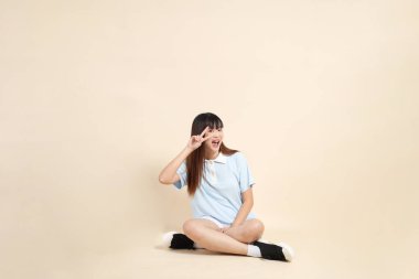 Açık mavi polo tişörtlü, beyaz şortlu, bej arka planda oturan genç Asyalı bir kadın. Üniversite öğrencisi, Gen Z, Summer