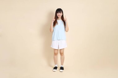 Açık mavi polo tişörtlü, beyaz şortlu genç bir Asyalı kadın bej bir arka planı işaret ediyor. Üniversite öğrencisi, Gen Z, Summer
