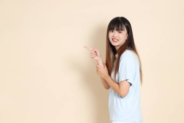 Açık mavi polo tişörtlü, beyaz şortlu genç bir Asyalı kadın bej bir arka planı işaret ediyor. Üniversite öğrencisi, Gen Z, Summer