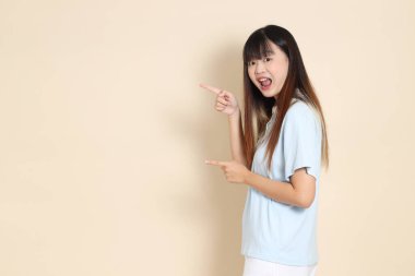 Açık mavi polo tişörtlü, beyaz şortlu genç bir Asyalı kadın bej bir arka planı işaret ediyor. Üniversite öğrencisi, Gen Z, Summer