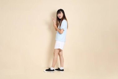 Açık mavi polo tişörtlü, beyaz şortlu genç bir Asyalı kadın bej bir arka planı işaret ediyor. Üniversite öğrencisi, Gen Z, Summer