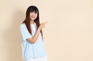 Açık mavi polo tişörtlü, beyaz şortlu genç bir Asyalı kadın bej bir arka planı işaret ediyor. Üniversite öğrencisi, Gen Z, Summer