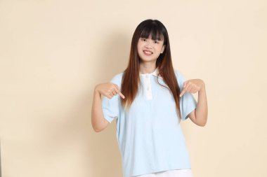 Açık mavi polo tişörtlü, beyaz şortlu genç bir Asyalı kadın bej bir arka planı işaret ediyor. Üniversite öğrencisi, Gen Z, Summer