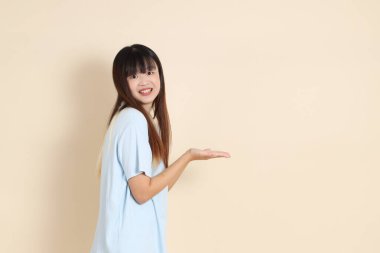 Açık mavi polo tişörtlü, beyaz şortlu genç bir Asyalı kadın bej bir arka planı işaret ediyor. Üniversite öğrencisi, Gen Z, Summer