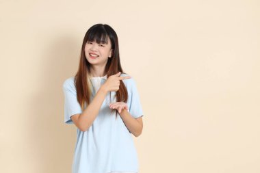 Açık mavi polo tişörtlü, beyaz şortlu genç bir Asyalı kadın bej bir arka planı işaret ediyor. Üniversite öğrencisi, Gen Z, Summer