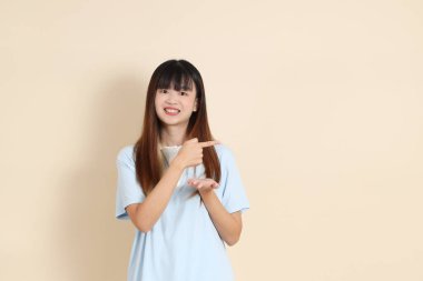 Açık mavi polo tişörtlü, beyaz şortlu genç bir Asyalı kadın bej bir arka planı işaret ediyor. Üniversite öğrencisi, Gen Z, Summer
