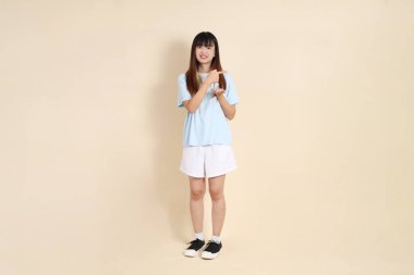 Açık mavi polo tişörtlü, beyaz şortlu genç bir Asyalı kadın bej bir arka planı işaret ediyor. Üniversite öğrencisi, Gen Z, Summer