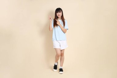 Açık mavi polo tişörtlü, beyaz şortlu genç bir Asyalı kadın bej bir arka planı işaret ediyor. Üniversite öğrencisi, Gen Z, Summer