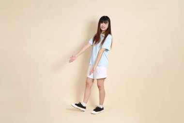 Açık mavi polo tişörtlü, beyaz şortlu genç bir Asyalı kadın bej bir arka planı işaret ediyor. Üniversite öğrencisi, Gen Z, Summer