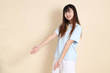 Açık mavi polo tişörtlü, beyaz şortlu genç bir Asyalı kadın bej bir arka planı işaret ediyor. Üniversite öğrencisi, Gen Z, Summer