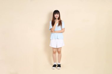 Açık mavi polo tişörtlü, beyaz şortlu genç bir Asyalı kadın bej bir arka planı işaret ediyor. Üniversite öğrencisi, Gen Z, Summer