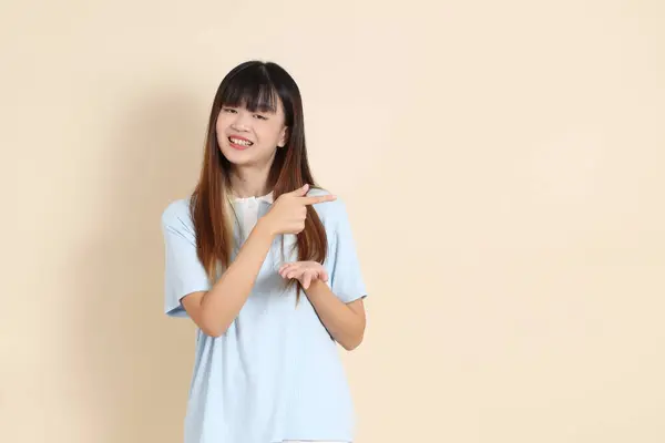 Açık mavi polo tişörtlü, beyaz şortlu genç bir Asyalı kadın bej bir arka planı işaret ediyor. Üniversite öğrencisi, Gen Z, Summer