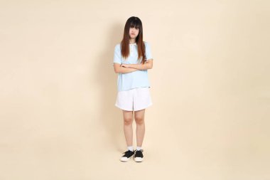 Açık mavi polo tişörtlü genç bir Asyalı kadın, ve beyaz şortlu, baş ağrısı ya da stresi olan düz bir bej arka plan. Üniversite öğrencisi, Gen Z, Summer