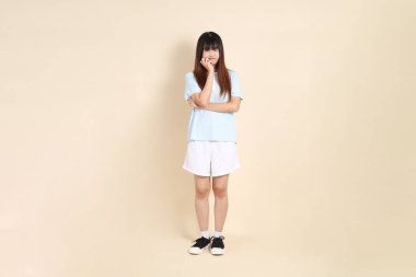 Açık mavi polo tişörtlü genç bir Asyalı kadın, ve beyaz şortlu, baş ağrısı ya da stresi olan düz bir bej arka plan. Üniversite öğrencisi, Gen Z, Summer