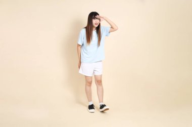 Açık mavi polo tişörtlü genç bir Asyalı kadın, ve beyaz şortlu, uzaktan bakan, düz bir bej arka plana bakan. Üniversite öğrencisi, Gen Z, Summer