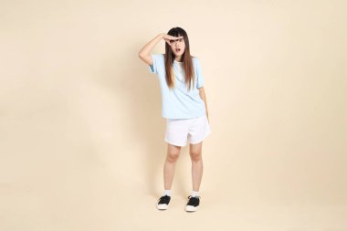 Açık mavi polo tişörtlü genç bir Asyalı kadın, ve beyaz şortlu, uzaktan bakan, düz bir bej arka plana bakan. Üniversite öğrencisi, Gen Z, Summer