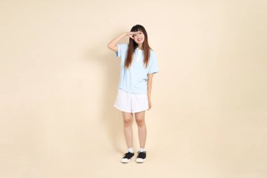 Açık mavi polo tişörtlü genç bir Asyalı kadın, ve beyaz şortlu, uzaktan bakan, düz bir bej arka plana bakan. Üniversite öğrencisi, Gen Z, Summer