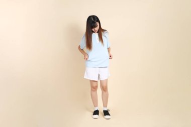 Açık mavi polo tişörtlü, beyaz şortlu genç Asyalı kadın bej arka planda giyiniyor. Üniversite öğrencisi, Gen Z, Summer