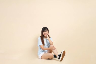 Açık mavi polo tişörtlü, beyaz şortlu, bej arka planda oturan genç Asyalı bir kadın. Üniversite öğrencisi, Gen Z, Summer