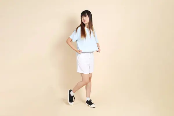 Açık mavi polo tişörtlü, beyaz şortlu genç Asyalı kadın bej arka planda giyiniyor. Üniversite öğrencisi, Gen Z, Summer