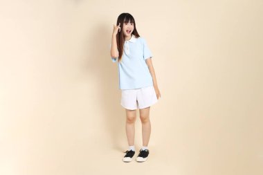 Açık mavi polo tişörtlü, beyaz şortlu genç bir Asyalı kadın bej arka planda sesleniyor. Üniversite öğrencisi, Gen Z, Summer