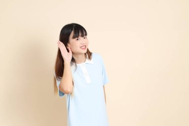 Açık mavi polo tişörtlü, beyaz şortlu, bej arka planda dinleyen ya da duyan genç Asyalı bir kadın. Üniversite öğrencisi, Gen Z, Summer