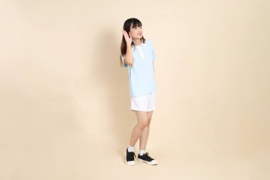 Açık mavi polo tişörtlü, beyaz şortlu, bej arka planda dinleyen ya da duyan genç Asyalı bir kadın. Üniversite öğrencisi, Gen Z, Summer