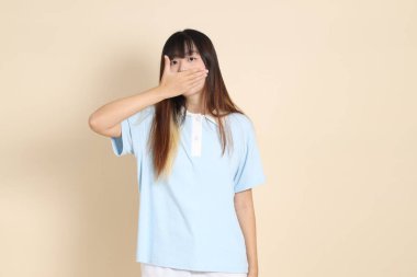 Açık mavi polo tişörtlü, beyaz şortlu, ağzı kapalı, bej arka planda sessiz bir jest yapan genç bir Asyalı kadın. Üniversite öğrencisi, Gen Z, Summer