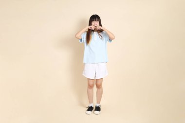 Açık mavi polo tişörtlü, beyaz şortlu, ağzı kapalı, bej arka planda sessiz bir jest yapan genç bir Asyalı kadın. Üniversite öğrencisi, Gen Z, Summer