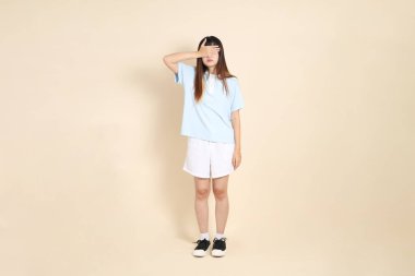Açık mavi polo tişörtlü, beyaz şortlu, bej arka planda gözleri olan genç Asyalı bir kadın. Üniversite öğrencisi, Gen Z, Summer