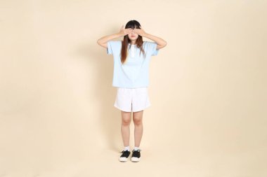 Açık mavi polo tişörtlü, beyaz şortlu, bej arka planda gözleri olan genç Asyalı bir kadın. Üniversite öğrencisi, Gen Z, Summer