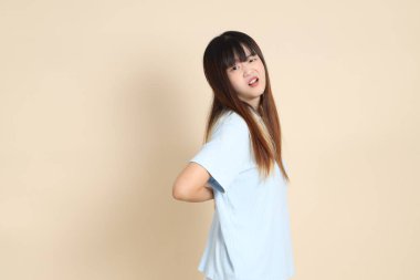 Açık mavi polo tişörtlü, beyaz şortlu, bej arka planda sırt ağrısı olan genç bir Asyalı kadın. Üniversite öğrencisi, Gen Z, Summer