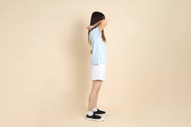 Açık mavi polo tişörtlü, beyaz şortlu, bej arka planda sırt ağrısı olan genç bir Asyalı kadın. Üniversite öğrencisi, Gen Z, Summer