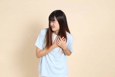 Açık mavi polo tişört ve beyaz şort giyen genç Asyalı bir kadın bej arka planda göğüs ağrısı çekiyor. Üniversite öğrencisi, Gen Z, Summer